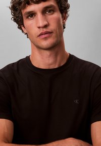 Ung mand med krøllet hår iført en ensfarvet sort kortærmet t-shirt med et lille "cK"-logo på brystet mod en grå baggrund.