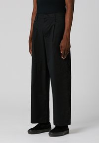 Pantalons larges noirs en tissu léger, avec une taille plate et des plis, et une texture lisse. Portés avec des baskets noires.