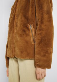 Veste en fausse fourrure marron avec une texture lisse, dotée d'une poche zippée et d'une finition en cuir tan le long de l'ouverture avant.