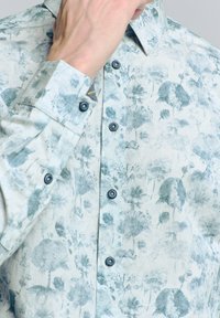 Lichtgekleurde blouse met knoopjes, voorzien van een bloemenpatroon in verschillende tinten groen. Gemaakt van een lichte stof, met contrasterende donkere knopen en kraagdetails.