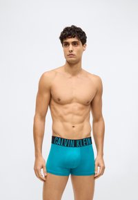 Pán bez trička s krátkými kudrnatými vlasy, oblečený v tyrkysových boxerkách Calvin Klein s černým páskem, stojící na jednobarevném světlém pozadí.