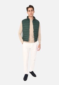 Gilet imbottito verde sopra una camicia a maniche lunghe beige, abbinato a pantaloni bianchi e scarpe da ginnastica blu navy. Tessuto lisci, senza motivi o hardware visibili.