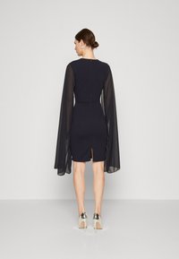 Κάλλυμα ναυτικού χρώματος με διαφανείς μακριές μανίκες, εφαρμ�οστή σιλουέτα, πίσω σκίσιμο και φερμουάρ στο λαιμό. Τα τακούνια με ασημί λεπτομέρειες ολοκληρώνουν την εμφάνιση.