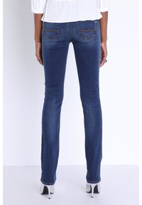 Dunkelblaue Jeans im Slim-Fit-Stil mit schrägen Gesäßtaschen und gelben Nähten. Das Model trägt silberne High-Heels.