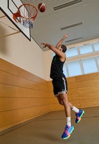 Un atleta in canotta nera senza maniche e pantaloni corti salta verso un canestro da basket, indossando sneakers dai colori vivaci con motivi intricati.