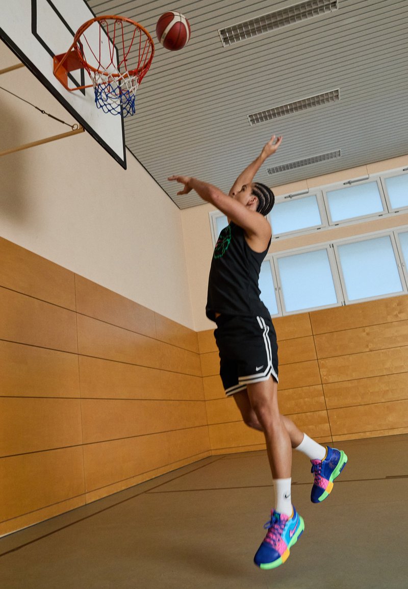 Athlet in einem schwarzen ärmellosen Trikot und Shorts springt in Richtung eines Basketballkorbs, trägt auffällig gefärbte Sneaker mit komplexen Mustern.