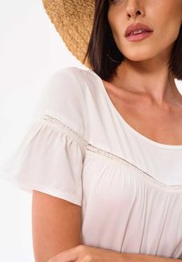 Lichtbeige blouse met korte afgeronde mouwen, voorzien van een kanten accent over de borst en een zachte, vloeiende snit.
