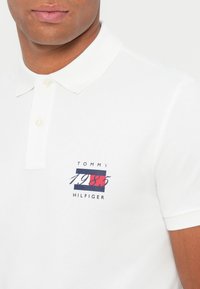 Valkoinen pikeepaita, joka on valmistettu teksturoidusta kankaasta ja jossa on kaulus ja kolme nappia. Etuosan design sisältää "TOMMY" ja "1985" sinisellä ja punaisella.