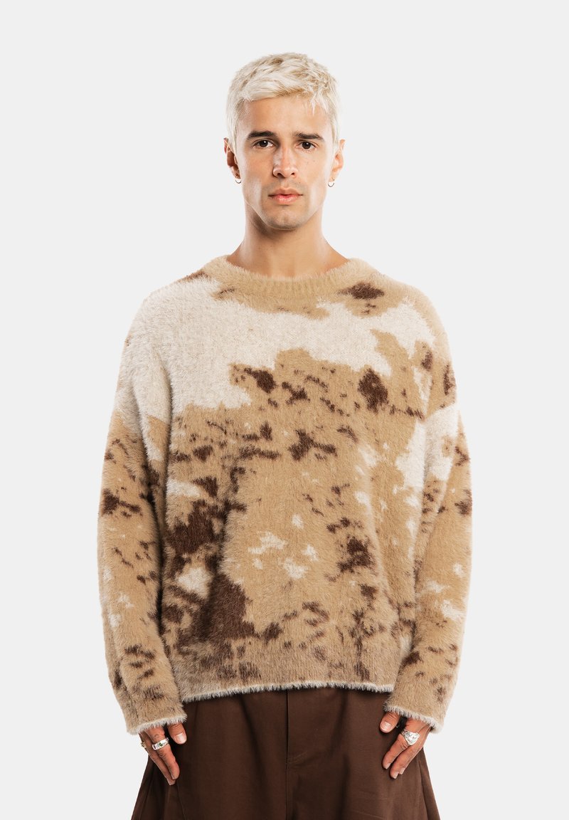 Jeune homme aux cheveux courts décolorés portant un pull duveteux beige et marron à motifs et un pantalon marron foncé, debout devant un fond uni.