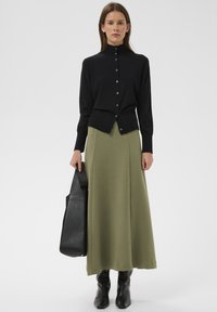 Cárdigan de punto negro con cuello alto y botones frontales, combinado con una falda larga de color verde oliva, complementado con un bolso tote negro y botines.