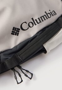 Κοντινό πλάνο τσάντας Columbia ανοιχτόγριζου και μαύρου χρώματος με εμφανές λογότυπο και διπλά φερμουάρ σε μαύρο λουρί.