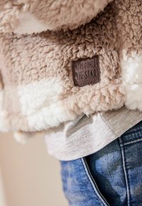 Uldet beige og hvidt plysjakke med en tekstureret overflade, der har et syet brunt mærke, der står "HER KOMMER STJERNEN."