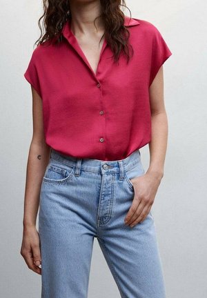 Kvinde iført en løstsiddende kortærmet rød bluse stoppet ned i lyseblå højtsiddende jeans, der står mod en ensfarvet baggrund.