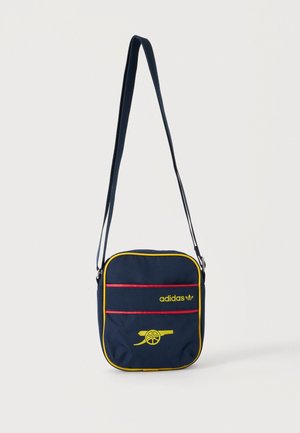 Μπλε ναυτικό crossbody τσάντα Adidas με κίτρινο φινίρισμα, κόκκινες λεπτομέρειες, λογότυπο Adidas και κίτρινο σχέδιο κανονιού στην μπροστινή τσέπη.