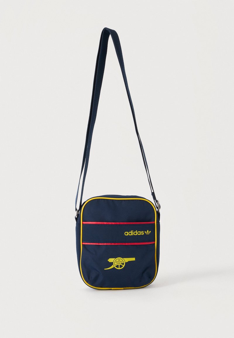Μπλε ναυτικό crossbody τσάντα Adidas με κίτρινο φινίρισμα, κόκκινες λεπτομέρειες, λογότυπο Adidas και κίτρινο σχέδιο κανονιού στην μπροστινή τσέπη.