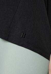 Detailní záběr na lem černého oděvu s malým vyšitým logem nad světle zelenou žebrovanou tkaninou.