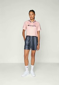 Camiseta rosa claro corta con el logo negro de "Champion", combinada con pantalones cortos a rayas gris oscuro y zapatillas blancas, mostrando un look deportivo y casual.