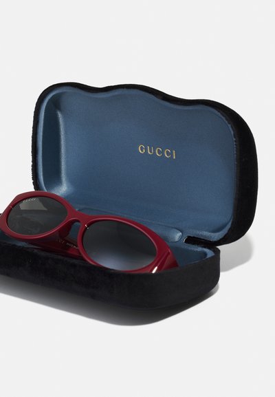 Punaiset muoviset aurinkolasit harmailla linsseillä mustassa, samettivuoratussa kotelossa, jossa on sininen vuori ja kultainen "GUCCI"-logo.