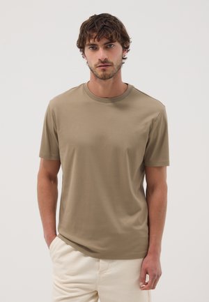 Mann mit kurzem lockigem braunen Haar, der ein schlichtes beige T-Shirt mit kurzen Ärmeln und weiße Hosen trägt und mit einer Hand in der Tasche steht.