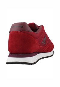 Zapatilla deportiva roja con parte superior de ante y textil, cuello acolchado, acentos morados en contraste y suela de goma blanca.