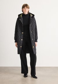 Gequilted zwarte lange jas met capuchon; voorzien van gouden hardware, geribde manchetten en een contrasterende patroonvoering. G gedragen over zwarte top en broek.