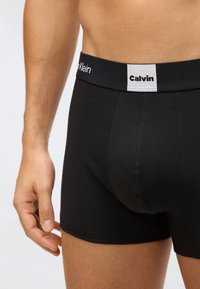 Detailní záběr na mužský trup a ruku, která má na sobě černé boxerky Calvin Klein s branded pasem.