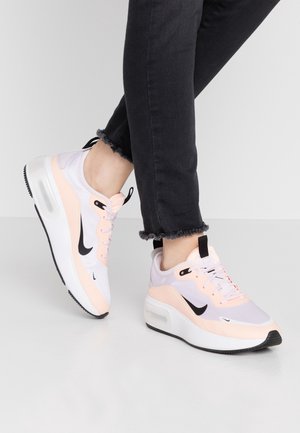 Osoba oblečená v černých kalhotách s roztřepeným lemem a bílých a broskvových teniskách Nike s viditelnými podešvemi s air-polštářky, pózující na světle šedé podlaze.