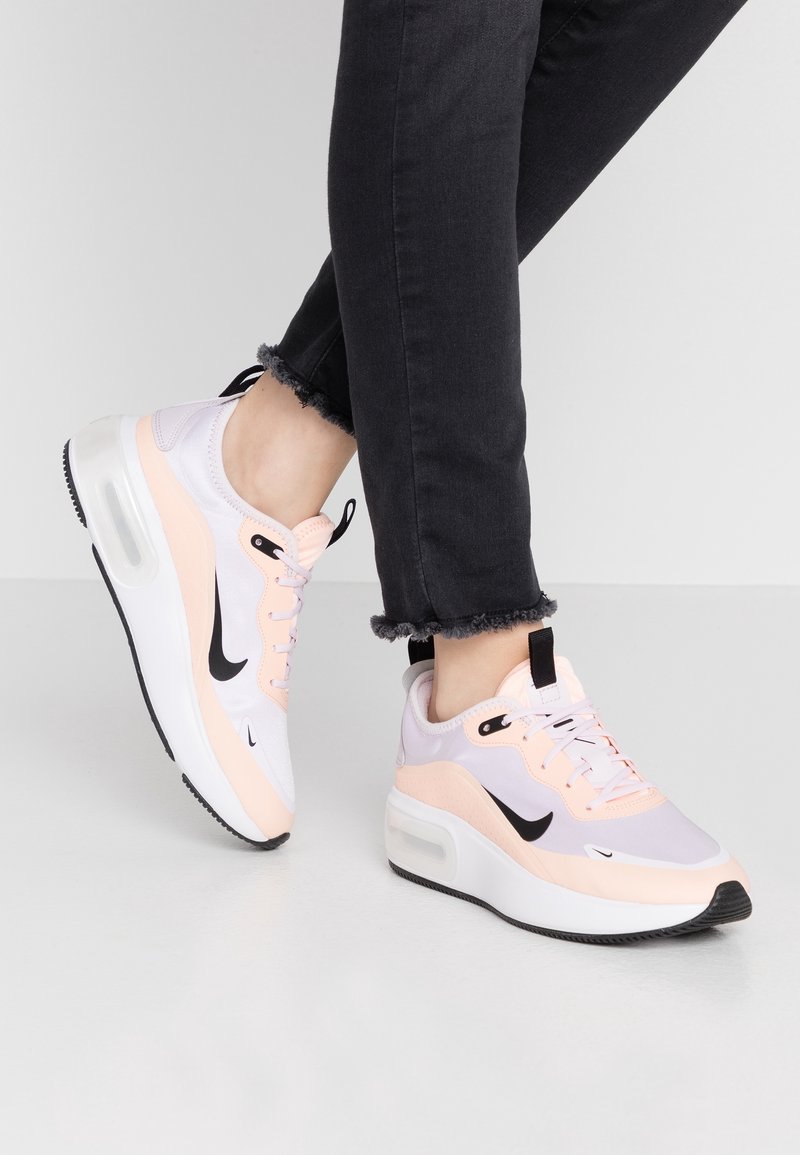 Personne portant un pantalon noir à ourlet effiloché et des baskets Nike blanches et pêche avec des semelles à coussin d'air visibles, posée sur un sol gris clair.