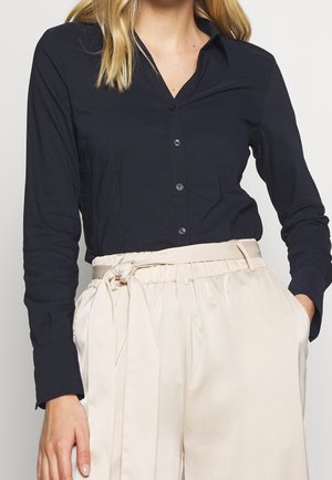 Navy long-sleeve button-up shirt with a collar, featuring a smooth texture and blue buttons, paired with light beige high-waisted trousers.