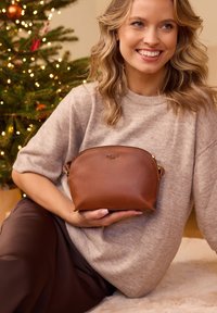 Braune Ledertasche mit rundem Oberteil, goldenen Beschlägen und Tragegriff an der Seite, gehalten vor einem neutralen Hintergrund mit Weihnachtsbaumschmuck.