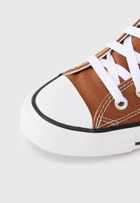 Καφέ πάνινο παπούτσι με λευκό λαστιχένιο κάλυμμα στα δάχτυλα, μαύρη επένδυση και λευκά κορδόνια. Διαθέτει υφασμάτινο επάνω μέρος με υφή και  μεταλλικά ρούντια για την εισαγωγή των κορδονιών.