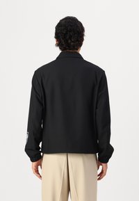Black jacket with a relaxed fit, featuring a collared neckline and elastic cuffs. Includes a patch detail on the left sleeve.