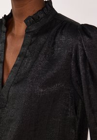 Black blouse featuring a ruffled collar, soft texture, and subtly shiny fabric. Tailored with puffed sleeves and a v-neckline.