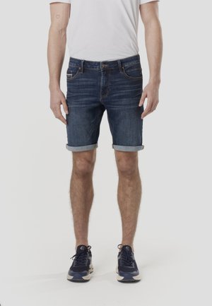 Szorty dżinsowe w ciemnym niebieskim kolorze z podwiniętymi nogawkami, posiadające pięć kieszeni i subtelne blaknięcie. Połączone z prostą białą koszulką i granatowymi trampkami.