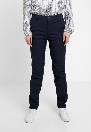 Persona che indossa pantaloni dritti blu navy, sneakers bianche e una camicetta a maniche lunghe con motivo, in piedi su sfondo bianco.