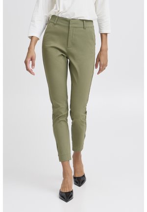 Femme portant un pantalon slim vert olive, un haut blanc à manches longues et des talons noirs à bout pointu, debout sur un fond blanc.