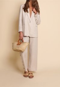 Kvinna iförd en beige linnekostym med knäppta byxor, hållande en vävd halmhandväska och bärande beige sandaler mot en beige bakgrund.