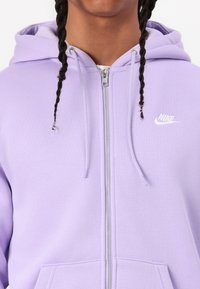 Fialová mikina Nike na zip s kapucí s nastavovací šňůrkou, předními kapsami a bílým logem na levé straně hrudi. Vyrobena z měkké, texturované látky.