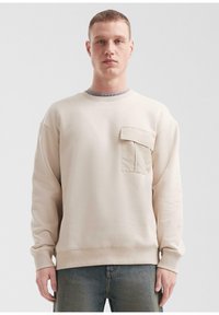 Młody mężczyzna z krótkimi włosami, ubrany w beżowy sweatshirt z kieszenią na klatce piersiowej i niebieskie dżinsy, stojący na jednorodnym tle.