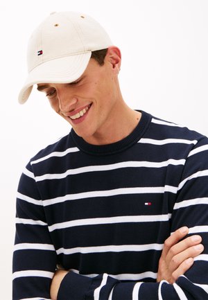 Glimlachende man met een witte pet en een marineblauw trui met witte horizontale strepen, armen gekruist, naar beneden kijkend.