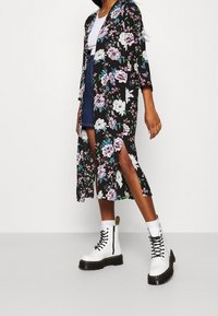 Blomstret kimono i sort stof med lilla og hvide blomster, lange ærmer og slids i siderne, parret med en hvid top og klodsede hvide støvler.