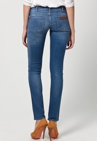 Smala blå denimjeans med mid-rise passform, två bakfickor och en brun logopatch på midjan. Bärs med bruna högklackade skor.