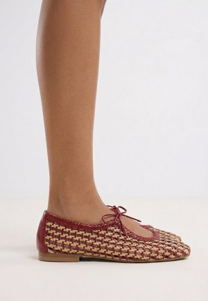 Pierna con zapato plano tejido en rojo y beige con detalle de lazo en la parte superior, mostrado sobre un fondo claro y sencillo.