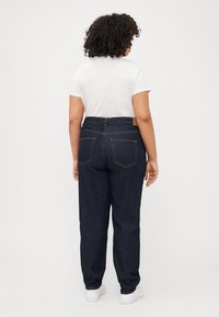 A person with curly hair wears a white shirt and dark jeans, viewed from the back, standing against a plain white background.