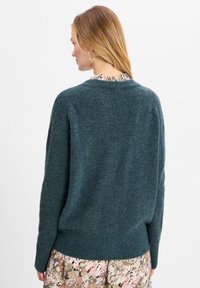 Teal wollen trui met geribde zoom en manchetten, voorzien van een ontspannen pasvorm en zachte textuur, zichtbaar vanaf de achterkant, gecombineerd met een bloemenrok.