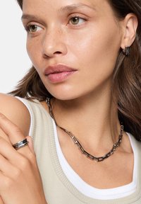 Bijoux en métal de couleur argent comprenant un collier à maillons rectangulaires et une bague assortie, associés à un haut beige texturé.