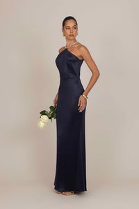 Navy satin mekko, jossa on epäsymmetrinen pääntie, istuva yläosa ja maahan ulottuva hame. Malli pitää kädessään valkoisia ruusuja ja käyttää kultaisia asusteita sekä sandaaleja.