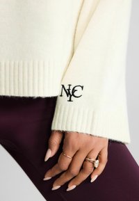Cremefarvet sweater med ribbede manchetter, prydet med sort broderet "NC" logo på ærmet. Hånd prydet med flere ringe og lange, spidse negle.