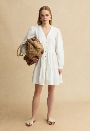 Giovane donna con vestito bianco abbottonato, che tiene una grande borsa intrecciata, indossa orecchini floreali e sandali aperti con dettagli a fiore.