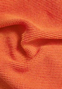 Textilstoff in lebhaftem Orange, mit einer strukturierten, geknoteten Webart, sichtbaren Nähten und einem weichen, leicht dehnbaren Gefühl.